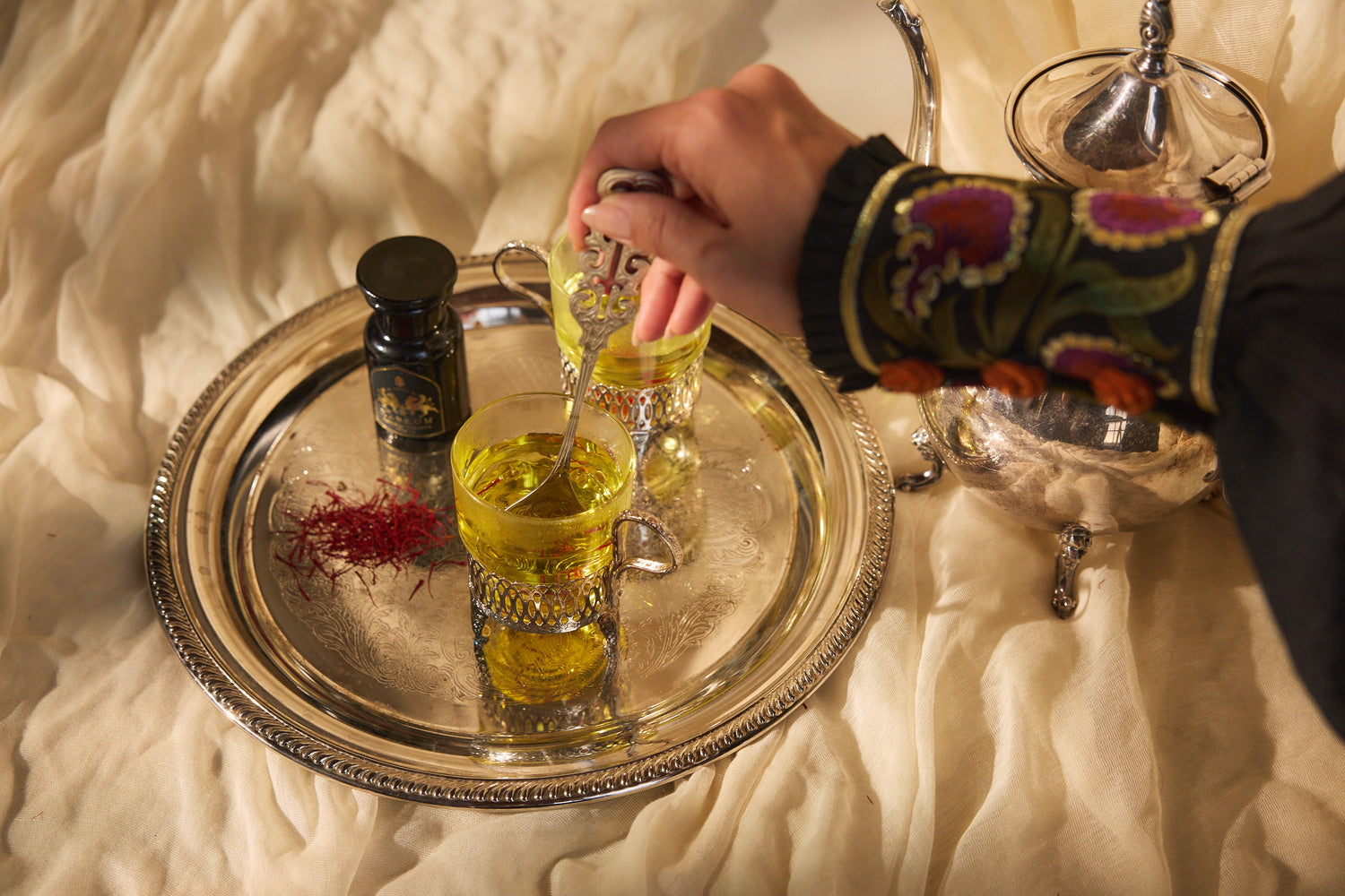 Person pouring a yellow liquid from a bottle into a glass on a silver tray with a textured surface.