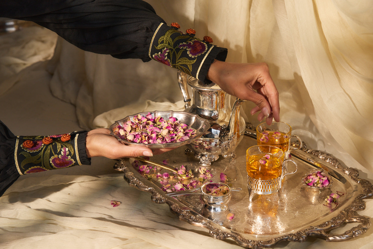 Person with embroidered sleeves serving Serene Soul botanical tea blend on a silver tray.